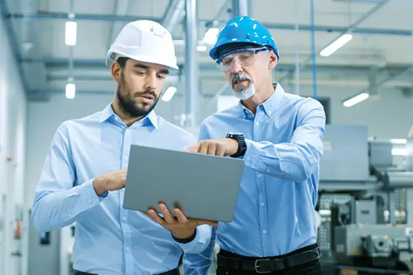 Two engineers wearing hard hats reviewing information on a laptop in a factory setting